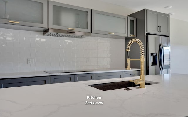 kitchen featuring light stone counters, black electric cooktop, gray cabinetry, stainless steel refrigerator with ice dispenser, and a sink