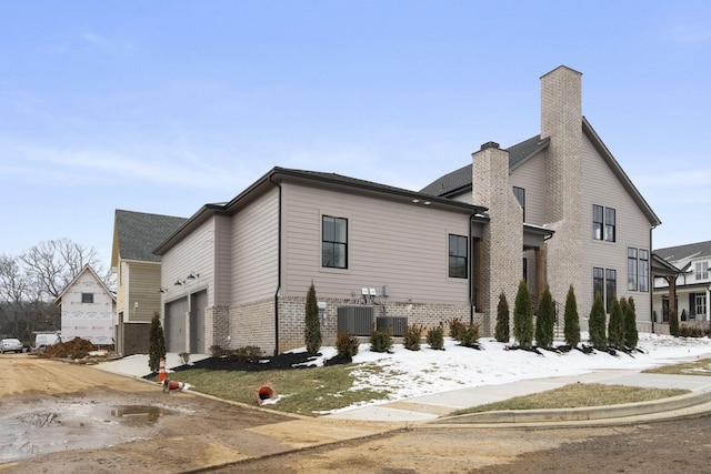 view of snow covered exterior featuring a garage, brick siding, a chimney, and central air condition unit