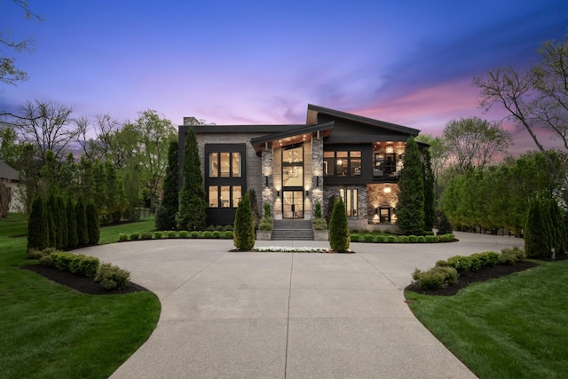 modern home featuring driveway, stone siding, a chimney, and a yard