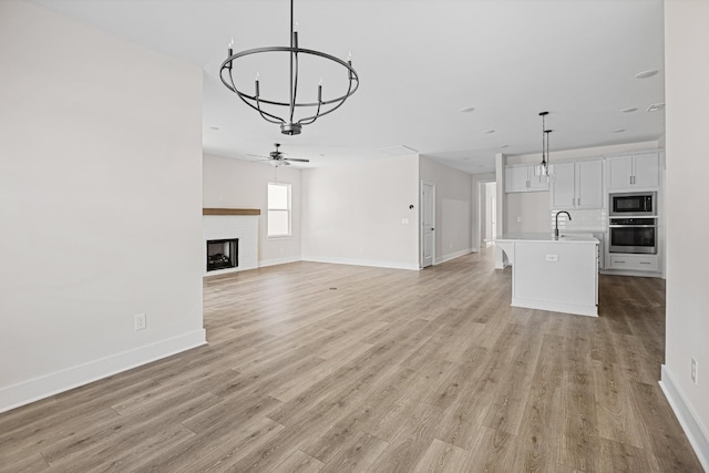 unfurnished living room with light wood-style flooring, ceiling fan with notable chandelier, a fireplace, a sink, and baseboards