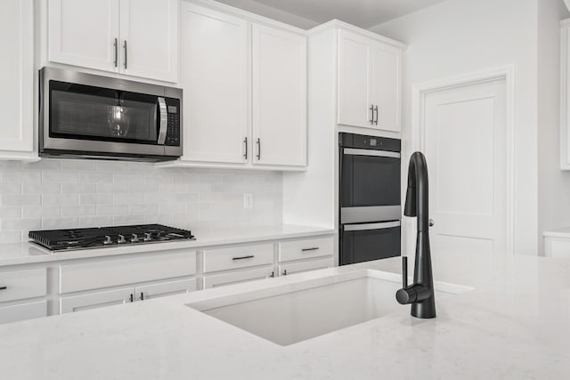 kitchen with stainless steel appliances, a sink, white cabinetry, backsplash, and light stone countertops