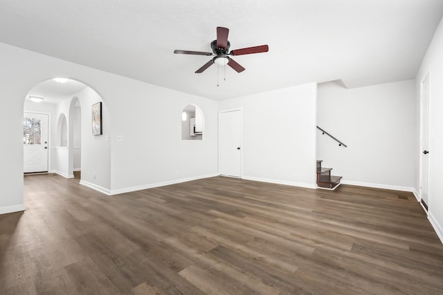 empty room with arched walkways, dark wood-style flooring, a ceiling fan, baseboards, and stairs