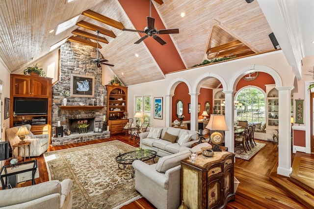 living room with arched walkways, a stone fireplace, wooden ceiling, dark wood-style floors, and ornate columns