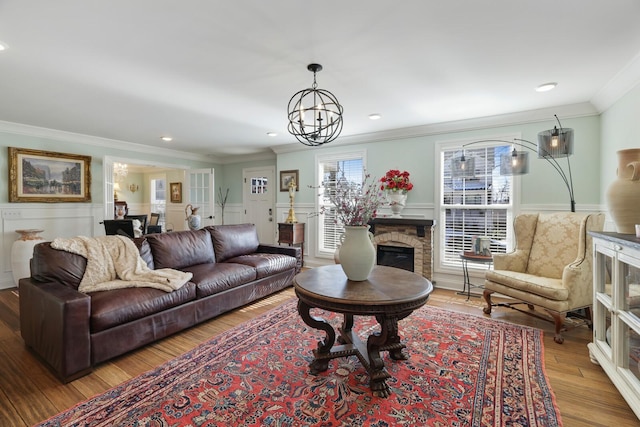 living area with wainscoting, light wood-style flooring, crown molding, a stone fireplace, and a chandelier