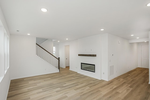 unfurnished living room featuring recessed lighting, light wood-style flooring, stairs, and a glass covered fireplace