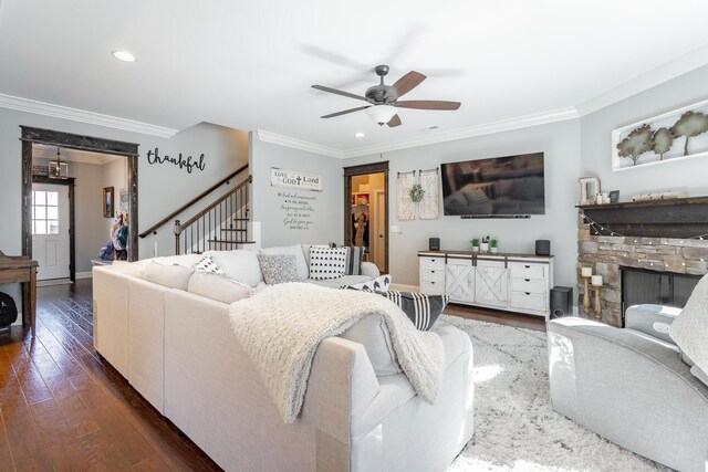 living room with a ceiling fan, dark wood-type flooring, stairs, crown molding, and a stone fireplace