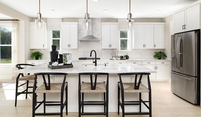 kitchen featuring hanging light fixtures, light countertops, stainless steel refrigerator with ice dispenser, wall chimney exhaust hood, and a center island with sink