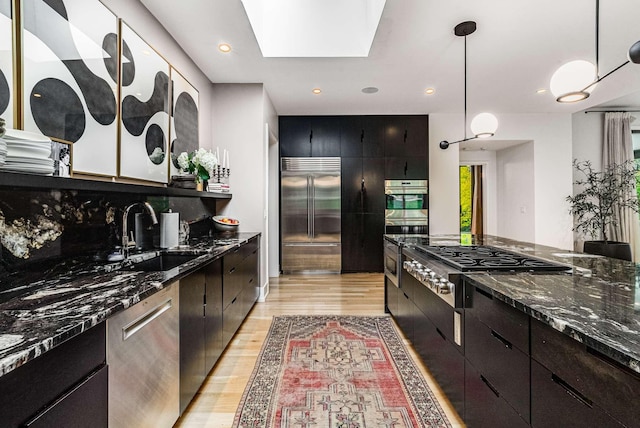 kitchen with stainless steel appliances, a sink, light wood-style floors, hanging light fixtures, and dark stone countertops