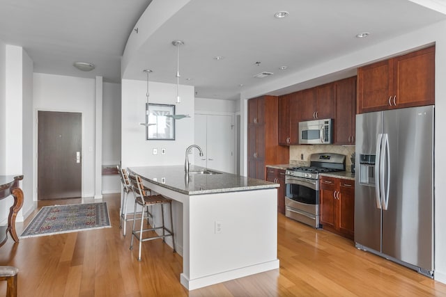 kitchen with stone countertops, a kitchen island with sink, stainless steel appliances, a sink, and light wood finished floors