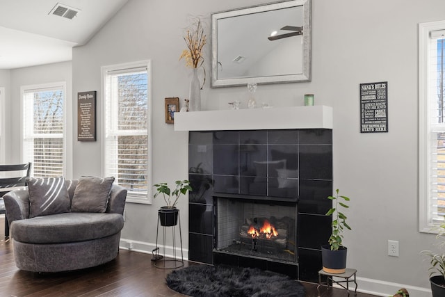 living room featuring dark wood-style flooring, visible vents, vaulted ceiling, a warm lit fireplace, and baseboards