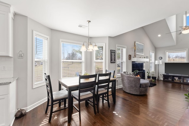 dining area with dark wood-style floors, vaulted ceiling, a warm lit fireplace, baseboards, and ceiling fan with notable chandelier