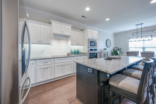 kitchen with a kitchen island with sink, stainless steel appliances, a sink, white cabinetry, and ornamental molding