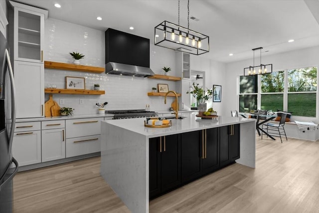 kitchen featuring decorative light fixtures, white cabinetry, wall chimney range hood, open shelves, and a center island with sink