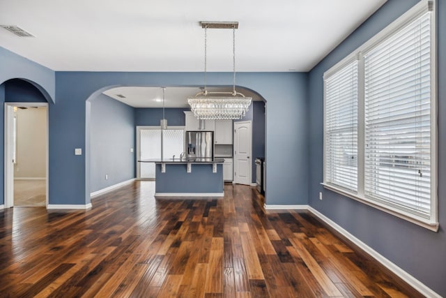 kitchen with a center island with sink, arched walkways, dark countertops, hanging light fixtures, and a kitchen bar