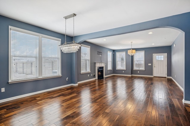 unfurnished living room featuring arched walkways, dark wood-style flooring, a notable chandelier, a warm lit fireplace, and baseboards