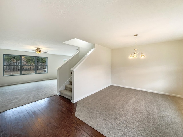 unfurnished living room with baseboards, stairway, dark colored carpet, and ceiling fan with notable chandelier