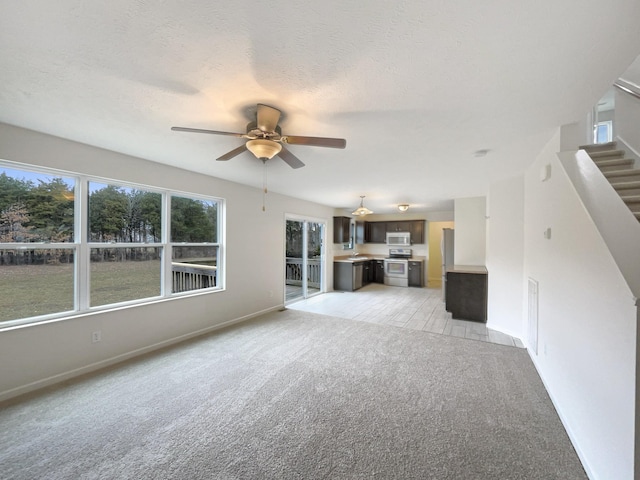 unfurnished living room featuring ceiling fan, a textured ceiling, light carpet, baseboards, and stairway