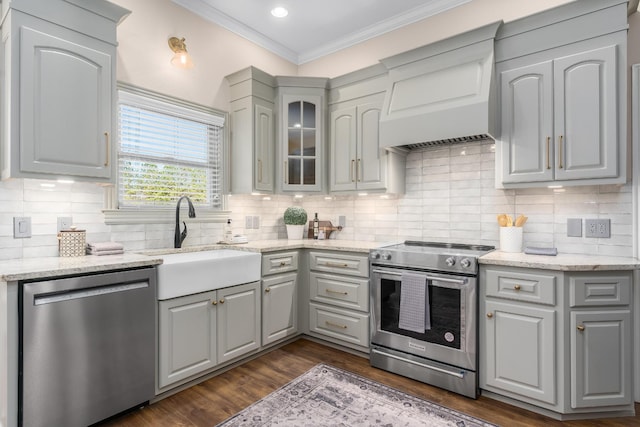 kitchen with gray cabinetry, stainless steel appliances, a sink, ornamental molding, and glass insert cabinets