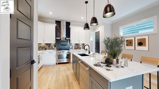 kitchen featuring a center island with sink, stainless steel appliances, hanging light fixtures, white cabinetry, and wall chimney range hood