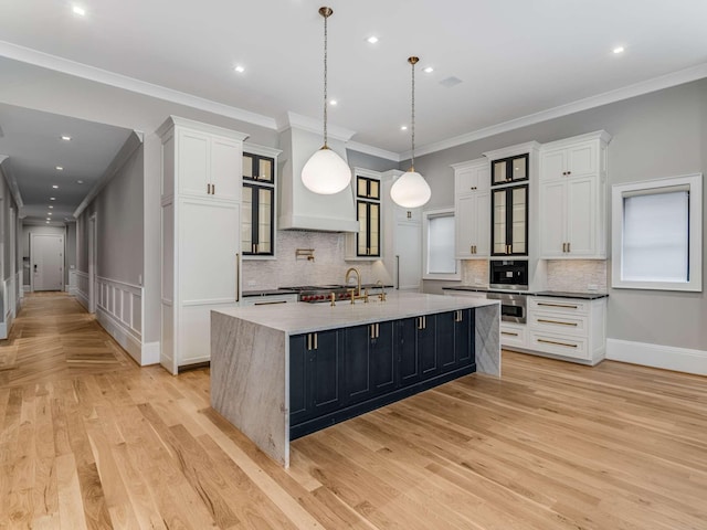 kitchen featuring decorative light fixtures, glass insert cabinets, white cabinets, light wood-type flooring, and a large island with sink