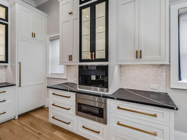 kitchen with white cabinetry, decorative backsplash, light wood finished floors, dark countertops, and glass insert cabinets