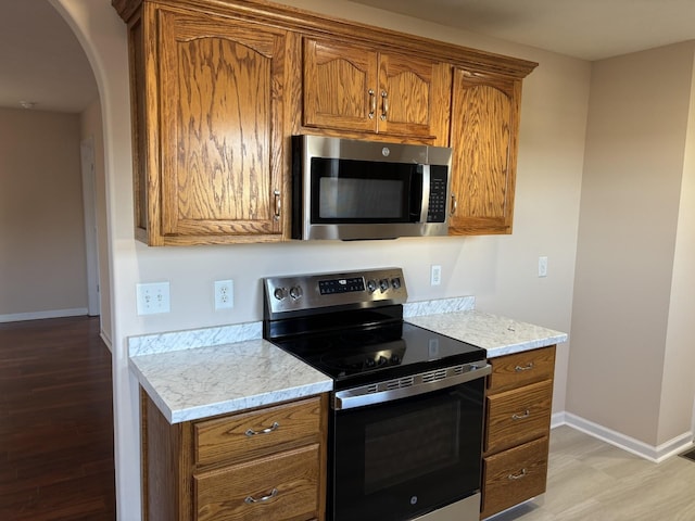 kitchen featuring arched walkways, brown cabinets, light wood finished floors, stainless steel appliances, and baseboards