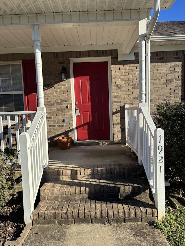 view of exterior entry with covered porch, brick siding, and roof with shingles