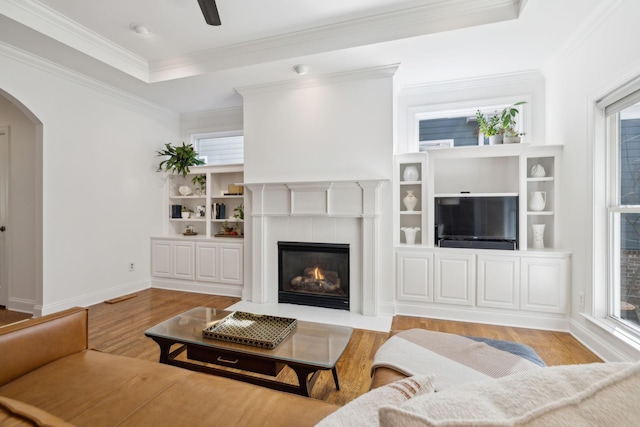 living room with arched walkways, crown molding, a raised ceiling, light wood-style flooring, and a fireplace with flush hearth