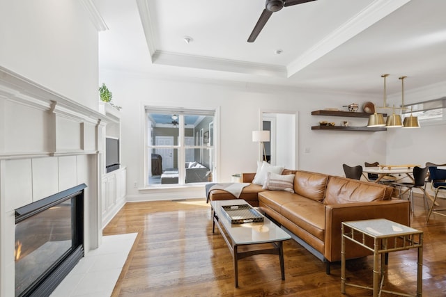 living room featuring a tile fireplace, a ceiling fan, ornamental molding, light wood-type flooring, and a raised ceiling