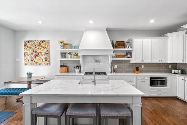 kitchen with custom range hood, stainless steel microwave, light stone countertops, a kitchen island with sink, and white cabinetry