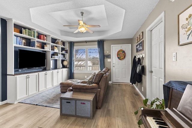 living area featuring light wood finished floors, a raised ceiling, a ceiling fan, a textured ceiling, and baseboards