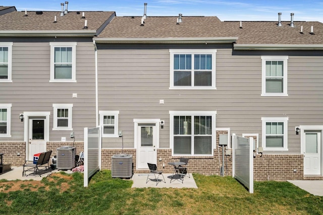 rear view of property featuring a shingled roof, a lawn, a patio, central air condition unit, and brick siding