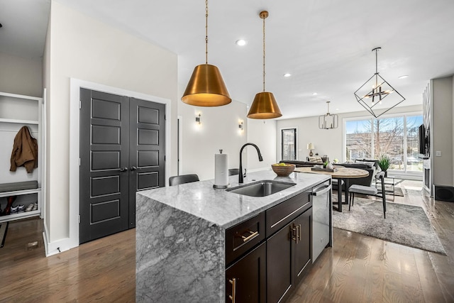 kitchen with pendant lighting, stainless steel dishwasher, a sink, an island with sink, and light stone countertops