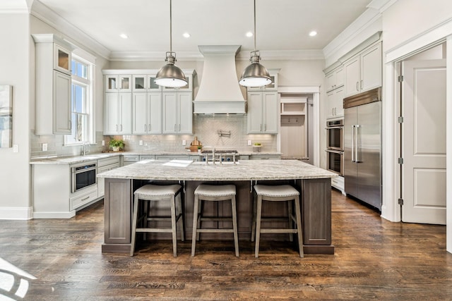 kitchen with hanging light fixtures, appliances with stainless steel finishes, a center island with sink, glass insert cabinets, and custom range hood