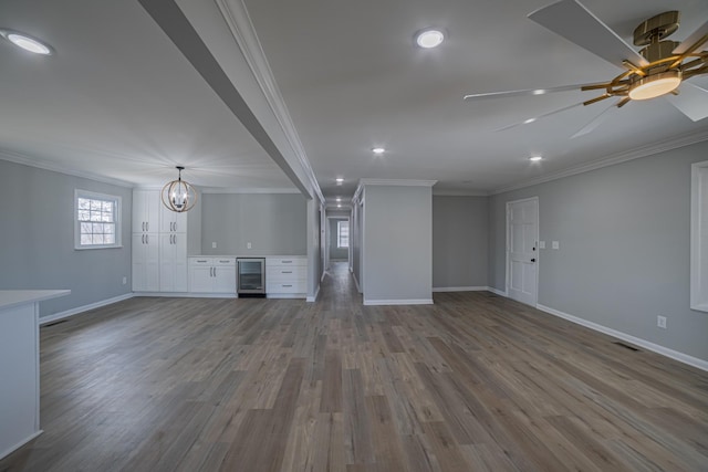 unfurnished living room featuring wine cooler, ceiling fan with notable chandelier, wood finished floors, baseboards, and crown molding