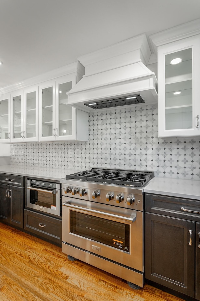 kitchen featuring stainless steel appliances, custom range hood, light countertops, and glass insert cabinets