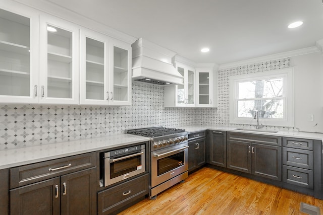 kitchen featuring glass insert cabinets, custom exhaust hood, stainless steel appliances, light countertops, and a sink