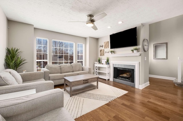 living area with baseboards, a tiled fireplace, dark wood-style floors, ceiling fan, and a textured ceiling