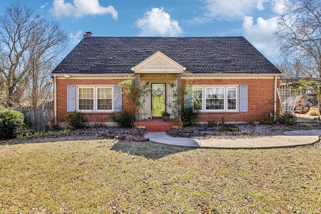 new england style home featuring brick siding, fence, roof with shingles, a chimney, and a front yard