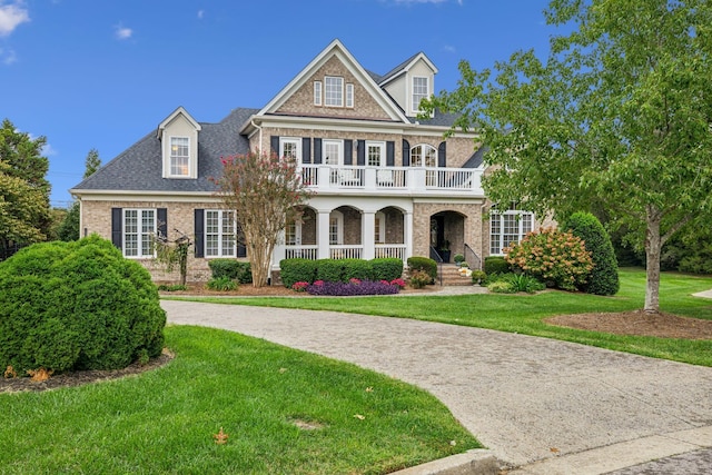 view of front facade featuring a porch, a balcony, brick siding, roof with shingles, and a front yard