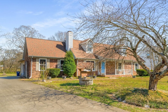 new england style home with a front lawn, driveway, covered porch, brick siding, and a chimney