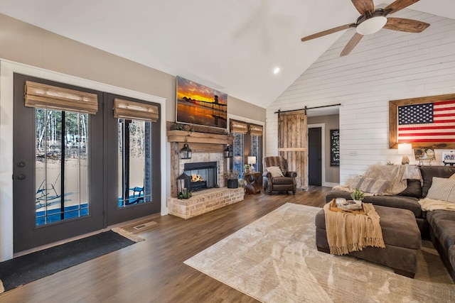 living room with high vaulted ceiling, a barn door, wood finished floors, a ceiling fan, and a brick fireplace