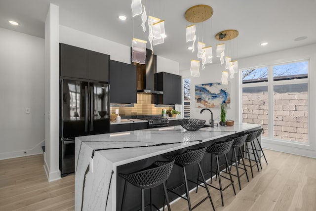 kitchen featuring a center island with sink, hanging light fixtures, decorative backsplash, wall chimney range hood, and black appliances