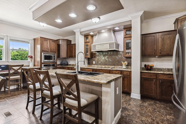 kitchen featuring a breakfast bar area, a kitchen island with sink, stainless steel appliances, premium range hood, and a sink