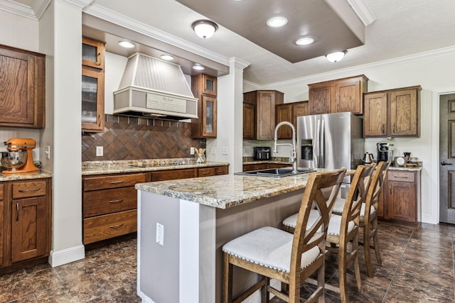 kitchen featuring a center island with sink, custom exhaust hood, glass insert cabinets, a sink, and light stone countertops