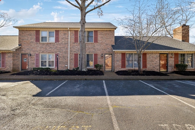 view of front facade with uncovered parking, brick siding, a chimney, and roof with shingles