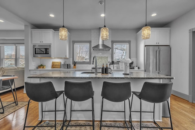 kitchen with a kitchen island with sink, white cabinets, appliances with stainless steel finishes, wall chimney range hood, and dark stone counters