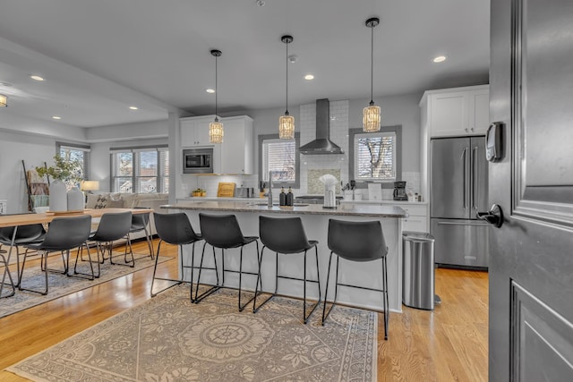 kitchen with stainless steel appliances, a kitchen island with sink, white cabinets, and wall chimney range hood