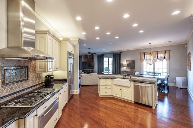 kitchen with cream cabinets, appliances with stainless steel finishes, open floor plan, a sink, and wall chimney range hood