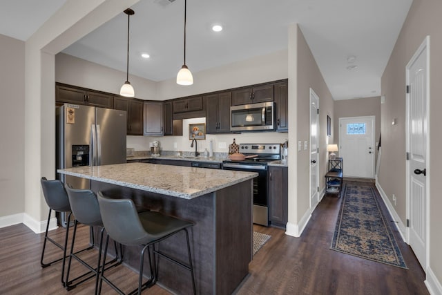 kitchen featuring dark brown cabinetry, appliances with stainless steel finishes, dark wood-type flooring, a center island, and light stone countertops